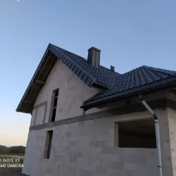 Niedokończony dom z czarnym dachem z dachówki ceramicznej, widoczne rynny i komin na tle błękitnego nieba o zmierzchu.