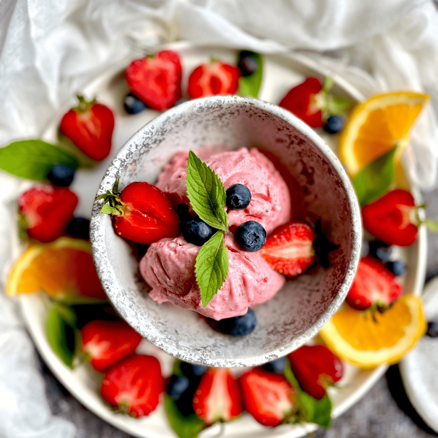 Różowe lody owocowe w miseczce, udekorowane borówkami i miętą, otoczone truskawkami i pomarańczą na białym talerzu.
