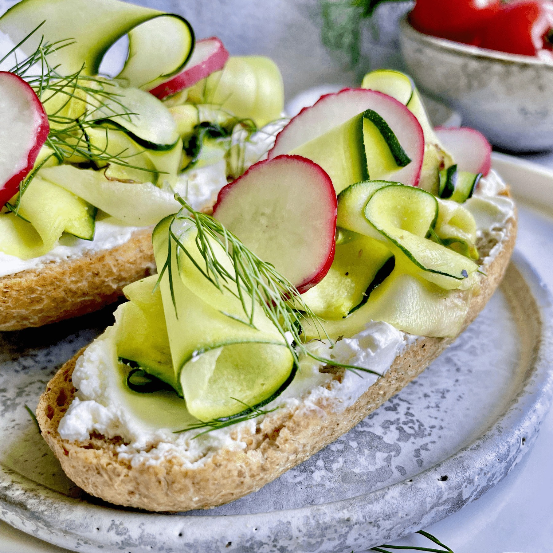 Dwie kanapki z twarożkiem, plasterkami rzodkiewki, ogórka i koperkiem na talerzu o marmurkowej fakturze.