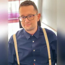 Portret uśmiechniętego adwokata w okularach i granatowej koszuli z szelkami, z widocznym fragmentem fioletowej zasłony w tle.