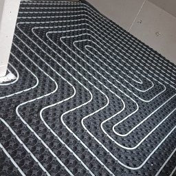 JK Instalacje - Białe rury ułożone w pętli na czarnej macie, tworzące system ogrzewania podłogowego w surowym wnętrzu poddasza.