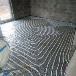 JK Instalacje - Montaż ogrzewania podłogowego: białe rury ułożone w pętle na metalowej siatce, zabezpieczone folią, widoczne czerwone mocowania, surowy stan ścian w tle.