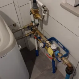 Złożony system rur i zaworów wodnych z licznikiem, zamontowany w łazience z białymi płytkami ściennymi i szarą podłogą, obok zmiękczacza wody.