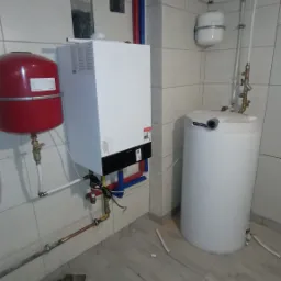 Biały piec grzewczy Viessmann z czerwonym zbiornikiem wyrównawczym, biały zasobnik ciepłej wody, rury instalacyjne w kolorach czerwonym i niebieskim, oraz fragment podłogi z płytek imitujących drewno.