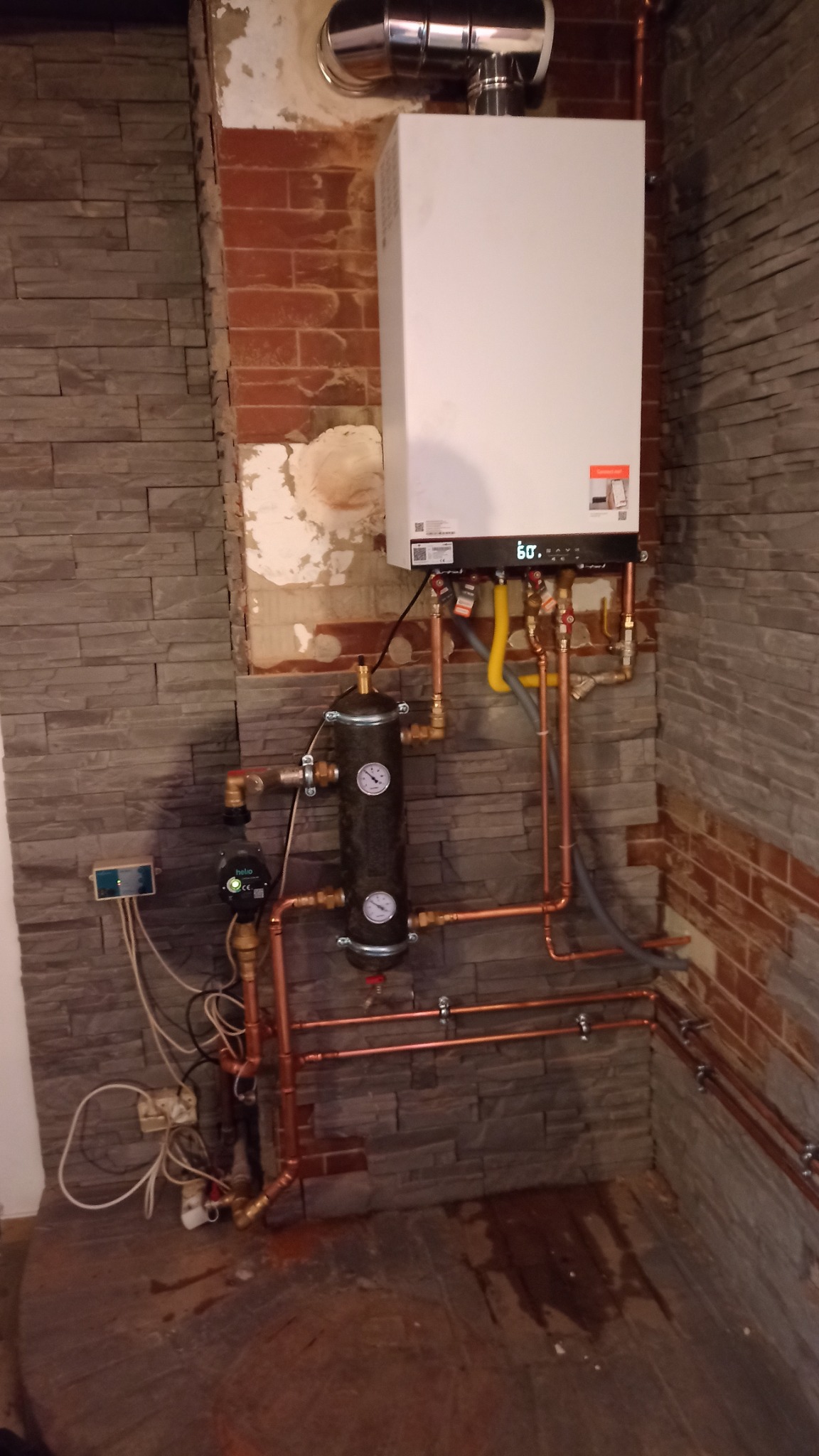 Nowoczesny piec gazowy kondensacyjny zamontowany na ścianie z dekoracyjną okładziną kamienną, widoczne miedziane rury instalacji, czarny zbiornik wyrównawczy z manometrami i elektroniczny sterownik...