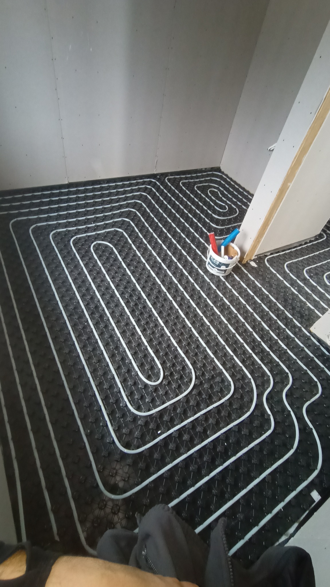 Montaż maty grzewczej z rurkami w kształcie pętli w narożnym pomieszczeniu z płytami kartonowo-gipsowymi, widoczne narzędzia w wiaderku.