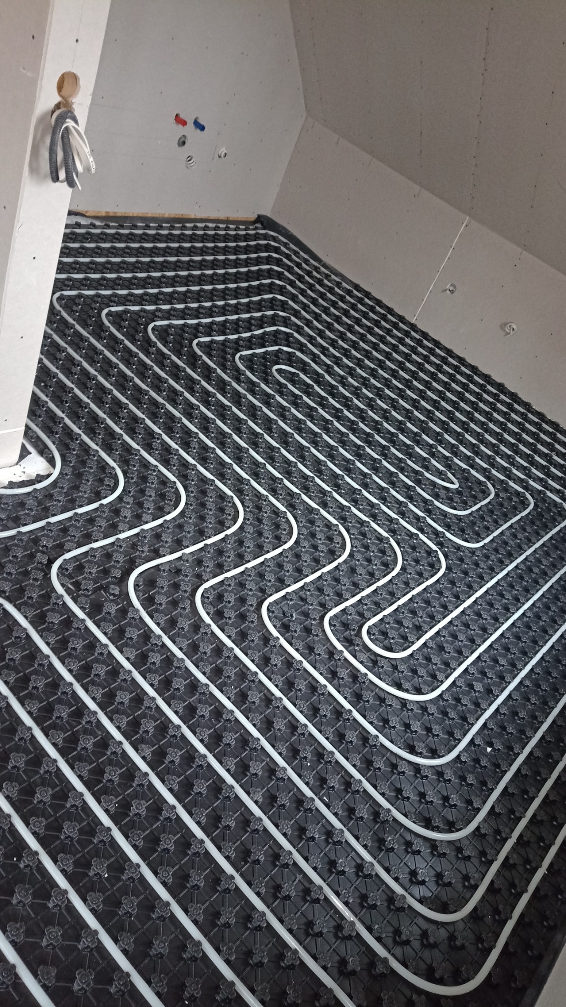 Białe rury ułożone w pętli na czarnej macie, tworzące system ogrzewania podłogowego w surowym wnętrzu poddasza.