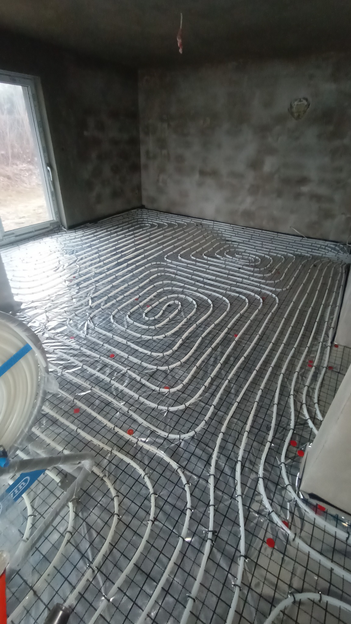 Montaż ogrzewania podłogowego: białe rury ułożone w pętle na metalowej siatce, zabezpieczone folią, widoczne czerwone mocowania, surowy stan ścian w tle.