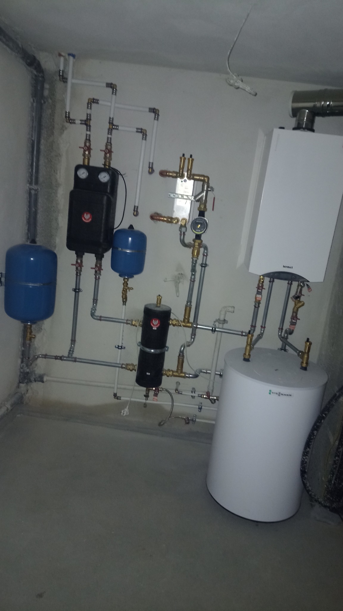Zainstalowany system grzewczy z kotłem wiszącym Termet, zbiornikiem Viessmann i dwoma niebieskimi naczyniami przeponowymi oraz czarnymi elementami armatury, widoczne rury i zawory.