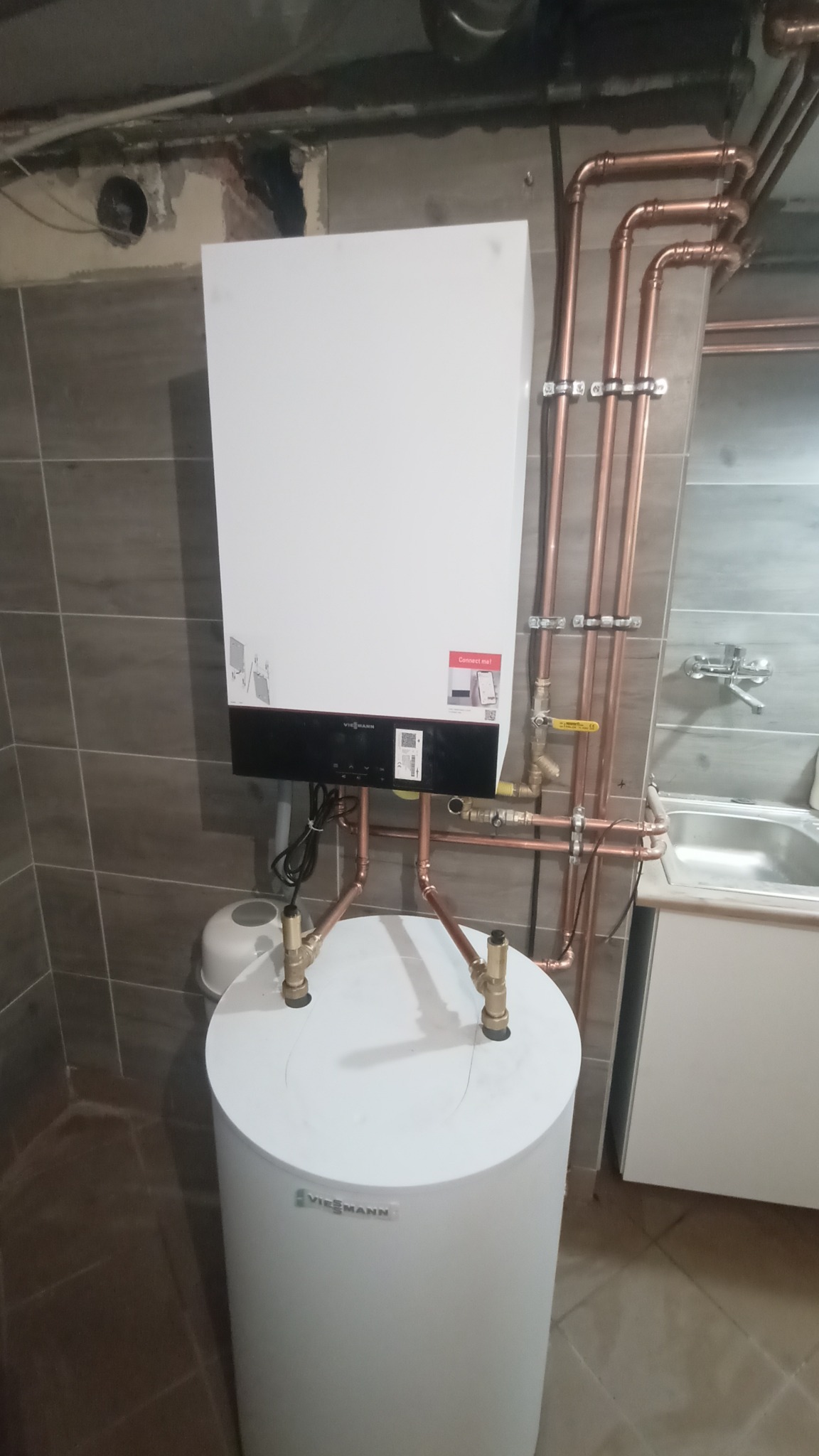 Nowoczesny, biały piec gazowy Viessmann zamontowany na białym zasobniku ciepłej wody, z widocznymi miedzianymi rurami instalacji, na tle szarych płytek ceramicznych.