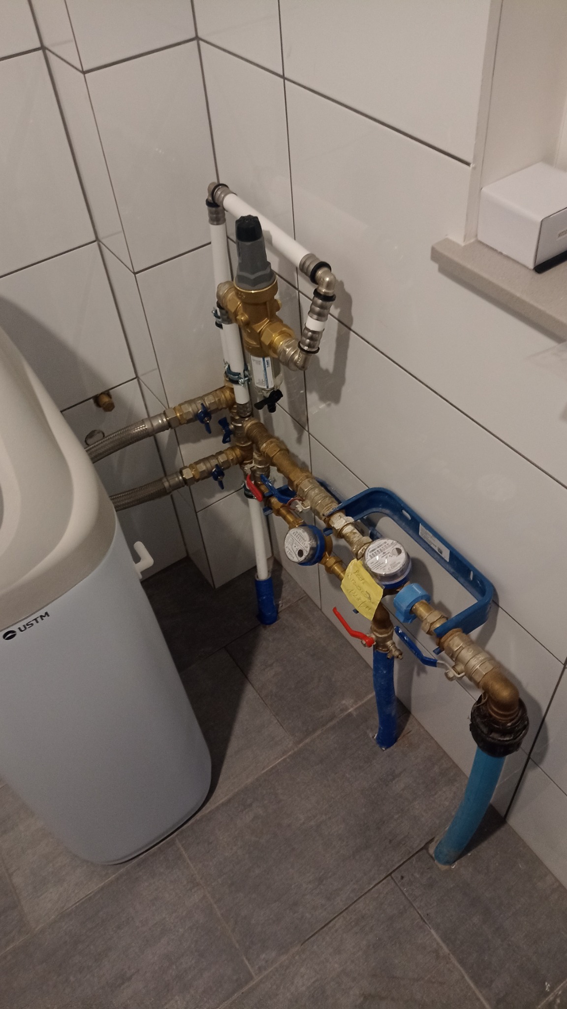 Złożony system rur i zaworów wodnych z licznikiem, zamontowany w łazience z białymi płytkami ściennymi i szarą podłogą, obok zmiękczacza wody.