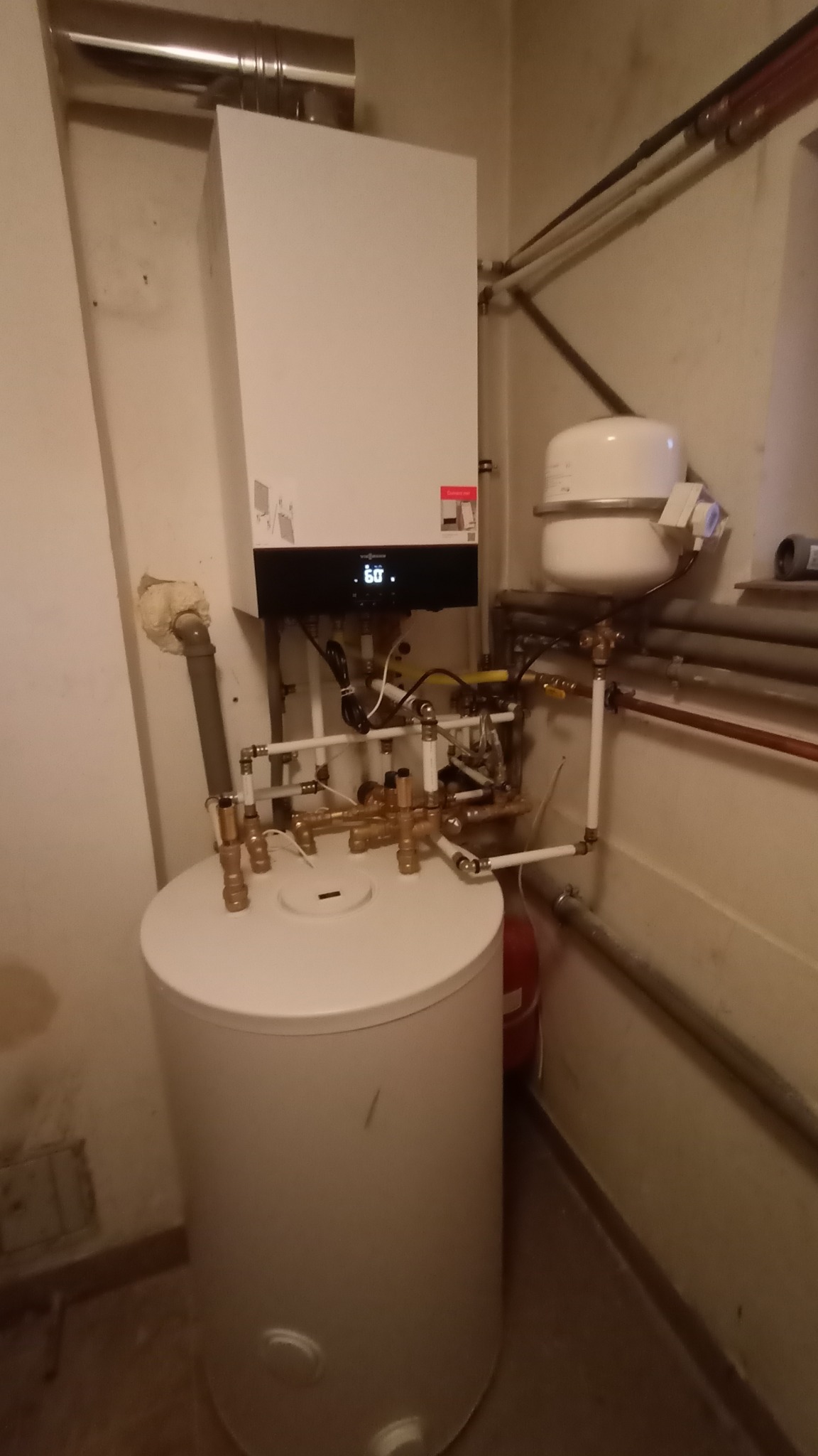 Biały kondensacyjny kocioł gazowy Viessmann z podłączonym zasobnikiem ciepłej wody i instalacją hydrauliczną, widoczne rury miedziane i plastikowe.