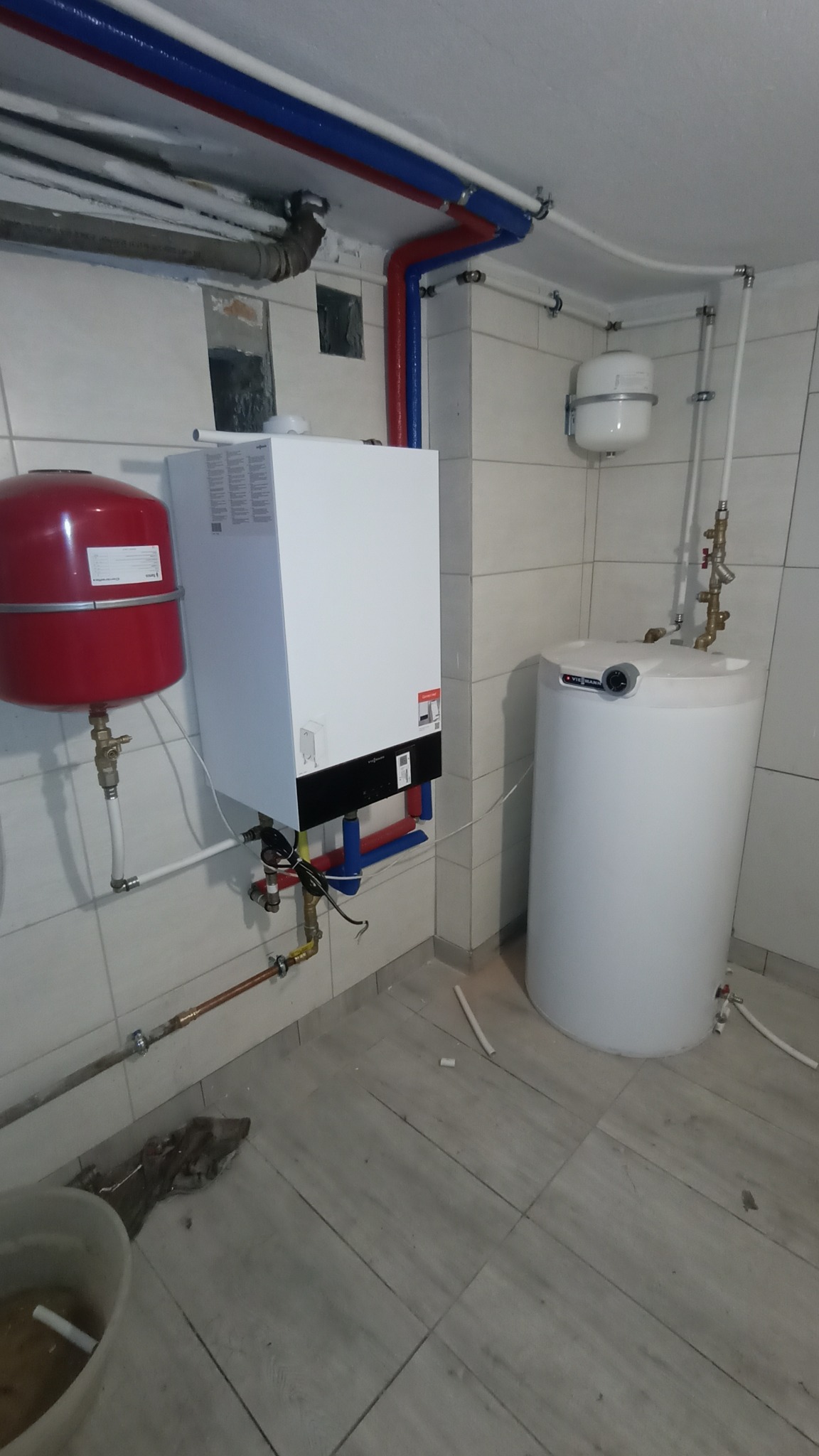 Biały piec grzewczy Viessmann z czerwonym zbiornikiem wyrównawczym, biały zasobnik ciepłej wody, rury instalacyjne w kolorach czerwonym i niebieskim, oraz fragment podłogi z płytek imitujących drewno.