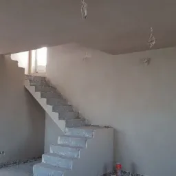 Wnętrze w trakcie remontu: świeżo otynkowane ściany, betonowe schody prowadzące na wyższą kondygnację, widoczne kable wystające z sufitu i ścian, surowy stan wykończenia.