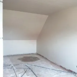 Pokój na poddaszu w trakcie remontu: ściany pokryte gładzią, widoczne instalacje elektryczne w podłodze, surowy stan wykończenia.