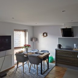 Patryk Sawicki - Jasne wnętrze z drewnianym stołem, szarymi krzesłami i elementami dekoracyjnymi, takimi jak wieniec na ścianie i poinsecja na stole. Widoczny fragment aneksu kuchennego z zabudową w odcieniach...