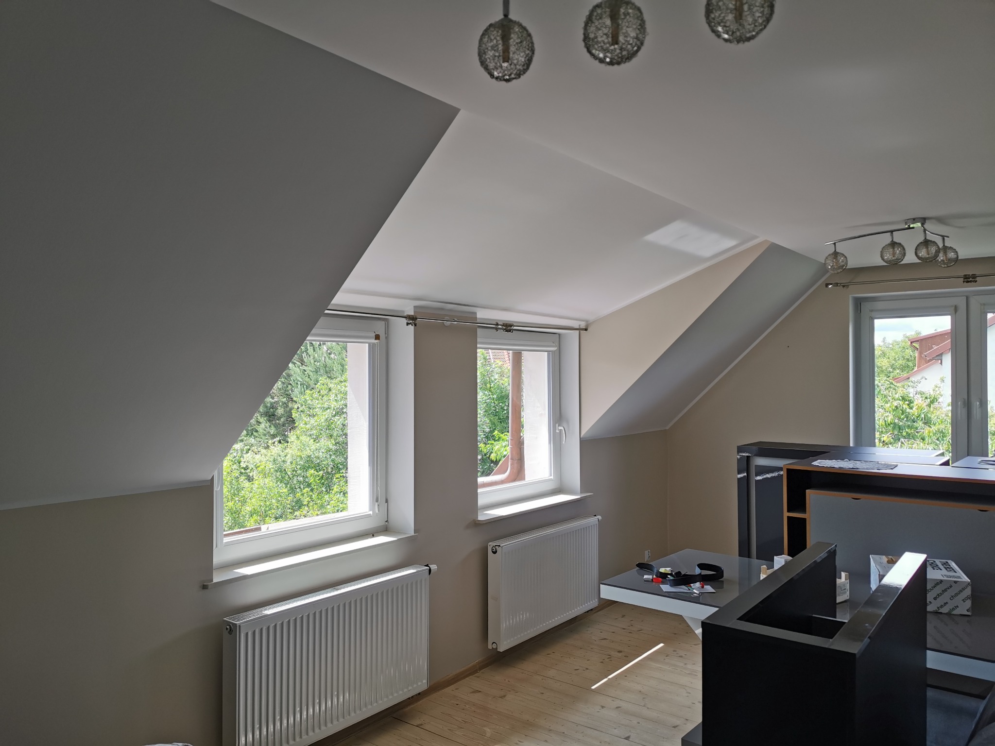 Jasne, przestronne poddasze w Gdańsku z dwoma oknami dachowymi, białymi grzejnikami i drewnianą podłogą. Ściany pomalowane na jasny odcień szarości, nowoczesne oświetlenie sufitowe.