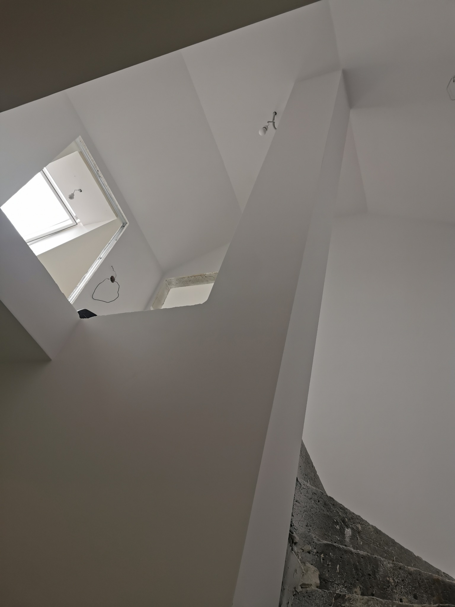Wnętrze w trakcie remontu: widok na białe ściany, sufit z oknem dachowym i fragment betonowych schodów w szarej tonacji.