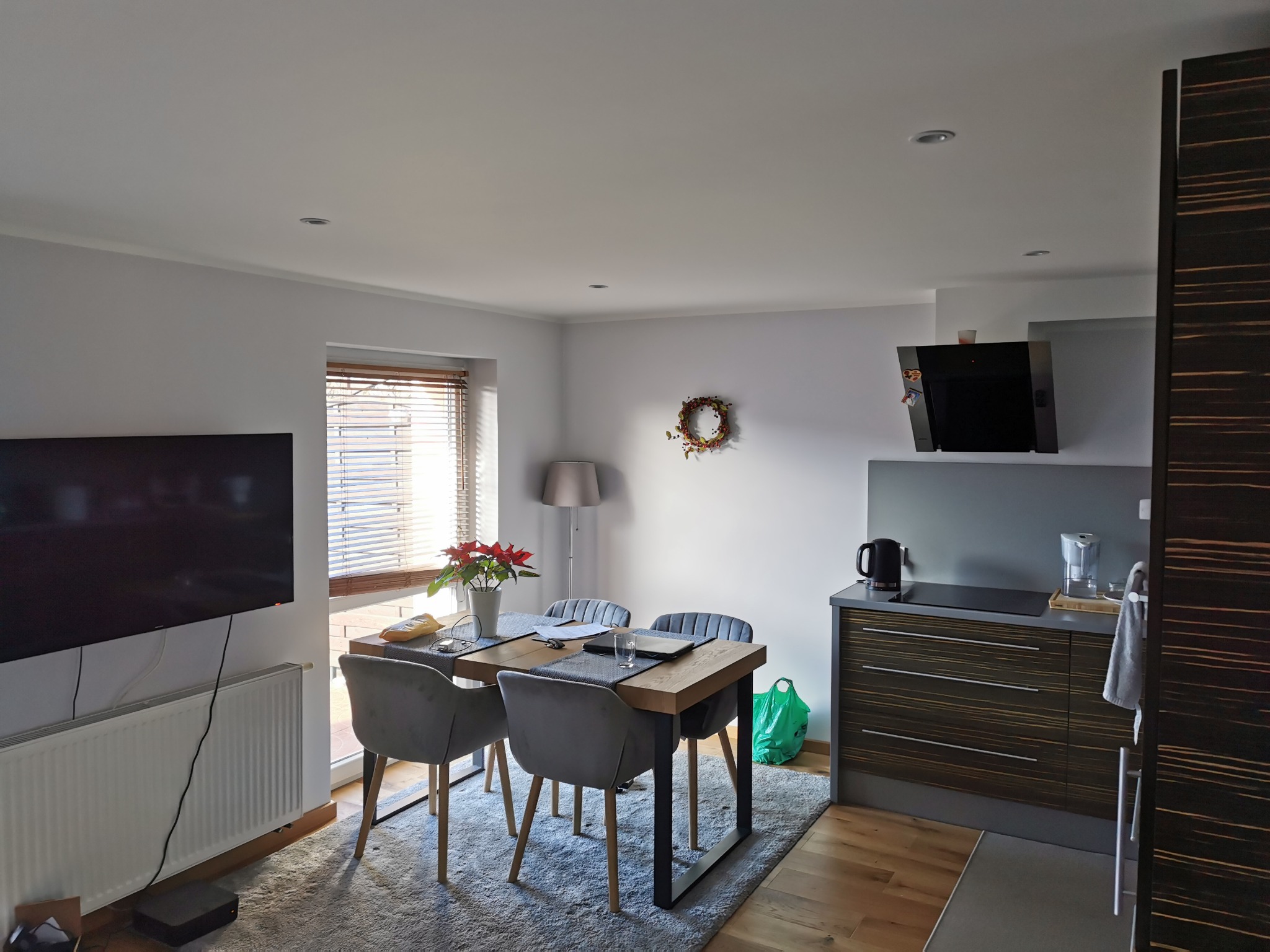 Jasne wnętrze z drewnianym stołem, szarymi krzesłami i elementami dekoracyjnymi, takimi jak wieniec na ścianie i poinsecja na stole. Widoczny fragment aneksu kuchennego z zabudową w odcieniach...