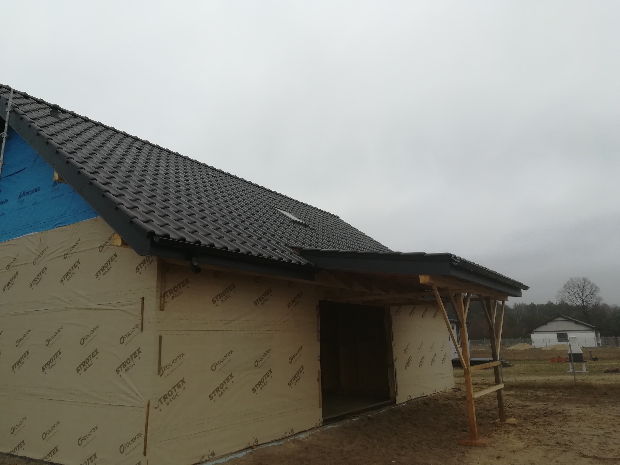 Dom w trakcie ocieplania poddasza, widoczna membrana izolacyjna StroTex Basic, dachówka ceramiczna Koramic, drewniane elementy konstrukcyjne i fragment ogrodu z domem w tle.