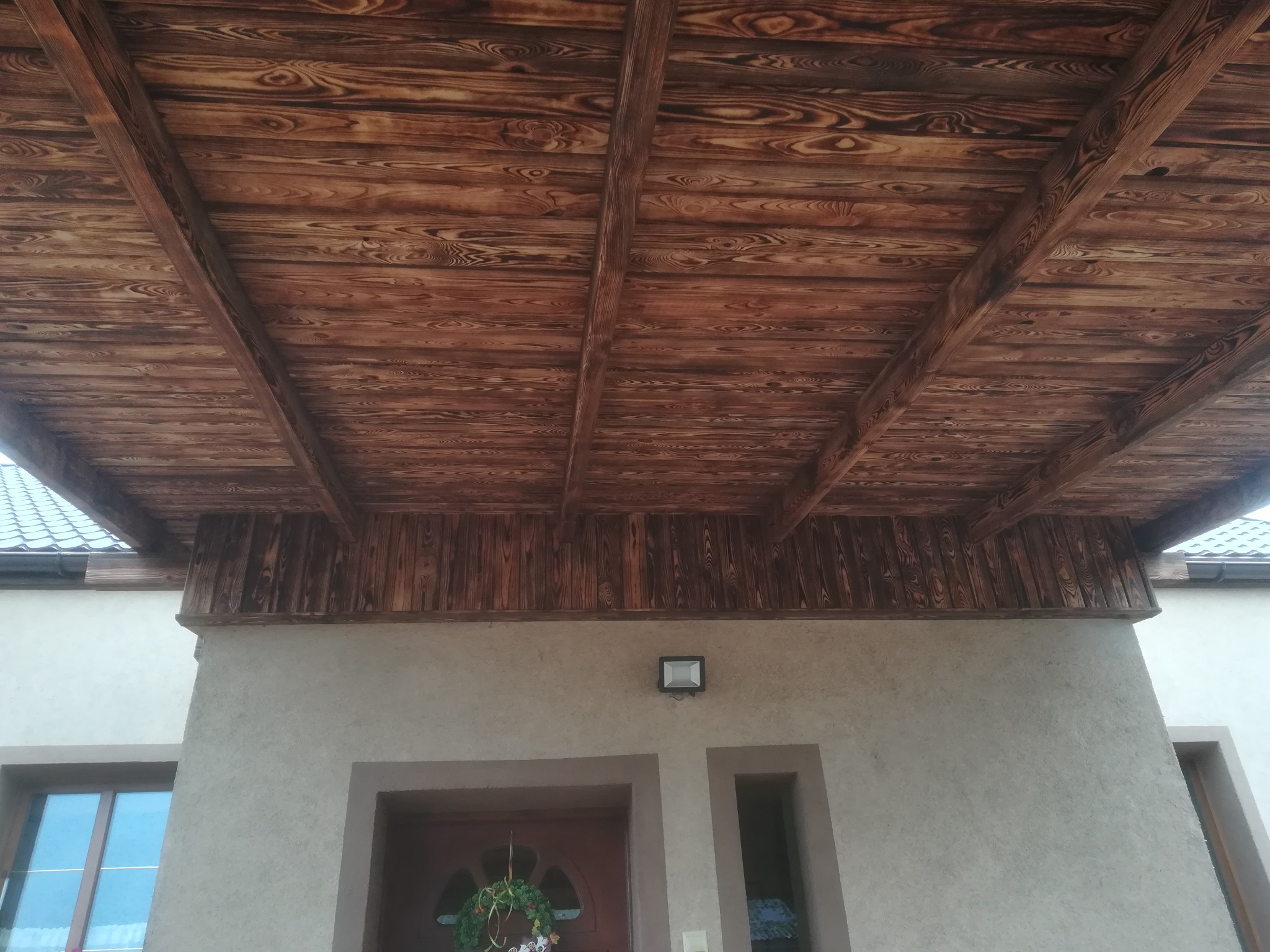 Drewniana boazeria podbitki dachowej z widocznym wzorem słojów, zamontowana na zewnątrz budynku, oświetlona lampą elewacyjną.