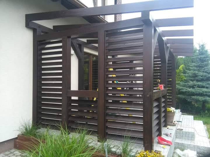 Drewniana pergola z żaluzjami przeciwsłonecznymi, pomalowana na ciemny brąz, przylegająca do białej ściany budynku; widoczne narzędzia i doniczki z kwiatami na schodach.