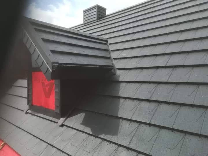 Szary dach z dachówki bitumicznej z kominem i oknem dachowym, zabezpieczonym czerwoną folią z nadrukiem.