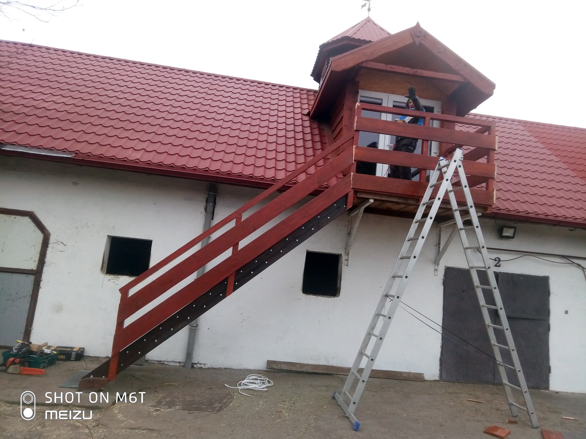 Wykończona zewnętrzna klatka schodowa z drewnianą balustradą prowadząca na balkon z małą nadbudówką na budynku z czerwoną dachówką, drabina oparta o balkon, narzędzia budowlane leżące obok schodów.