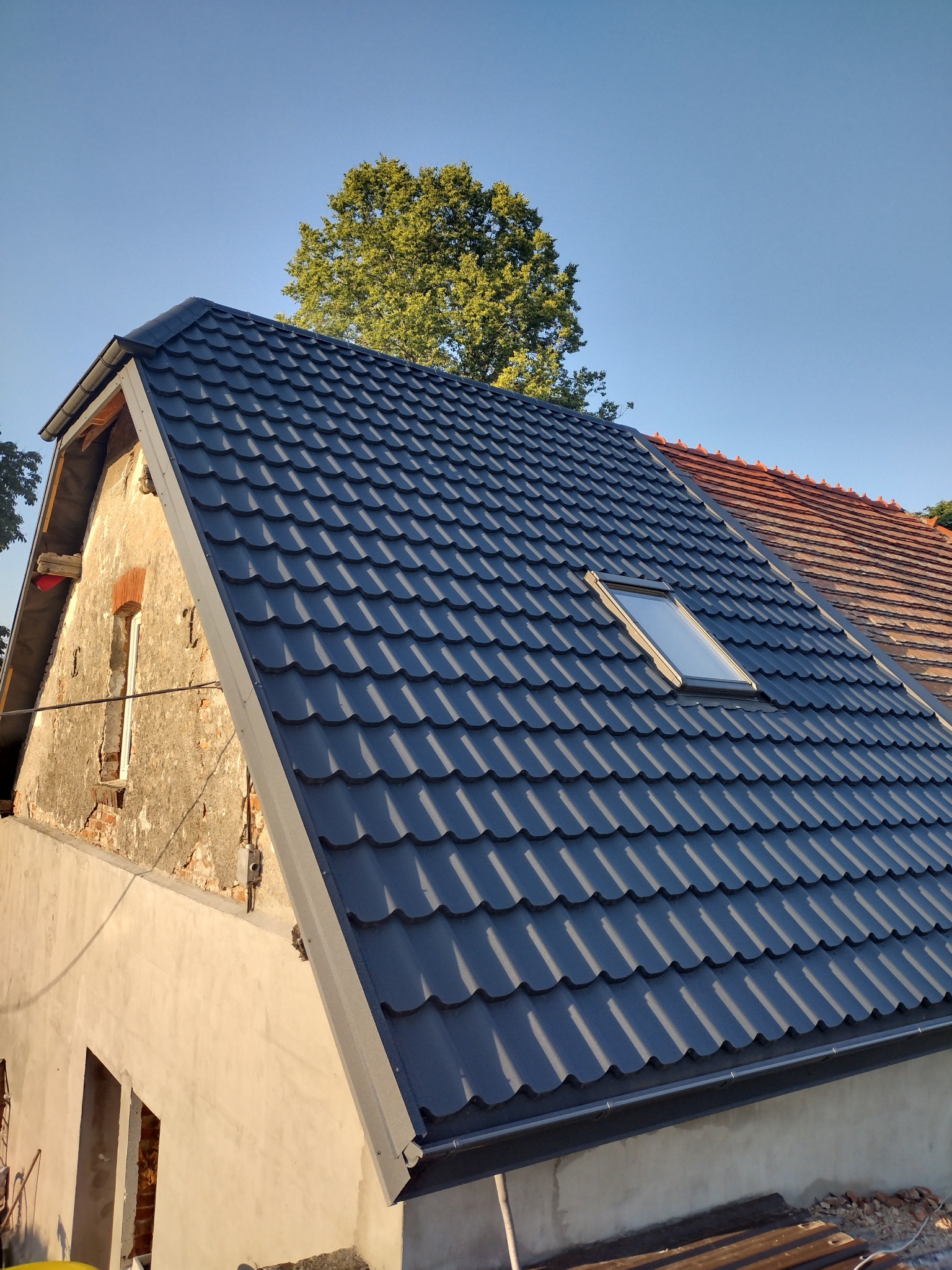 Częściowo odnowiony dach z ciemnoszarą blachodachówką i oknem dachowym, kontrastujący z fragmentem starej, czerwonej dachówki i nieotynkowaną ścianą szczytową.