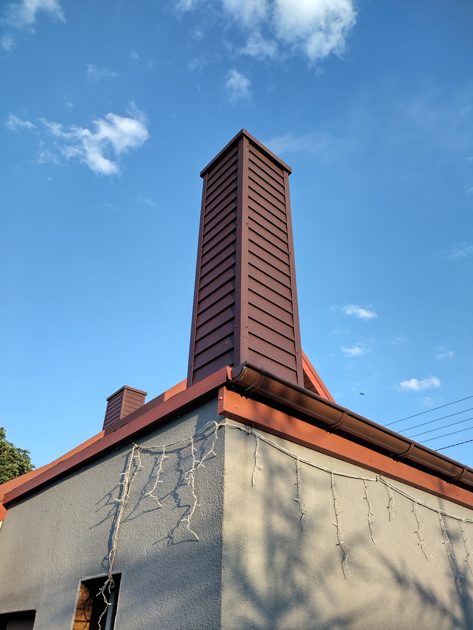 Widok z dołu na wysoki, brązowy komin z metalową obudową na szarym budynku z oknem w trakcie remontu, na tle błękitnego nieba z chmurami.