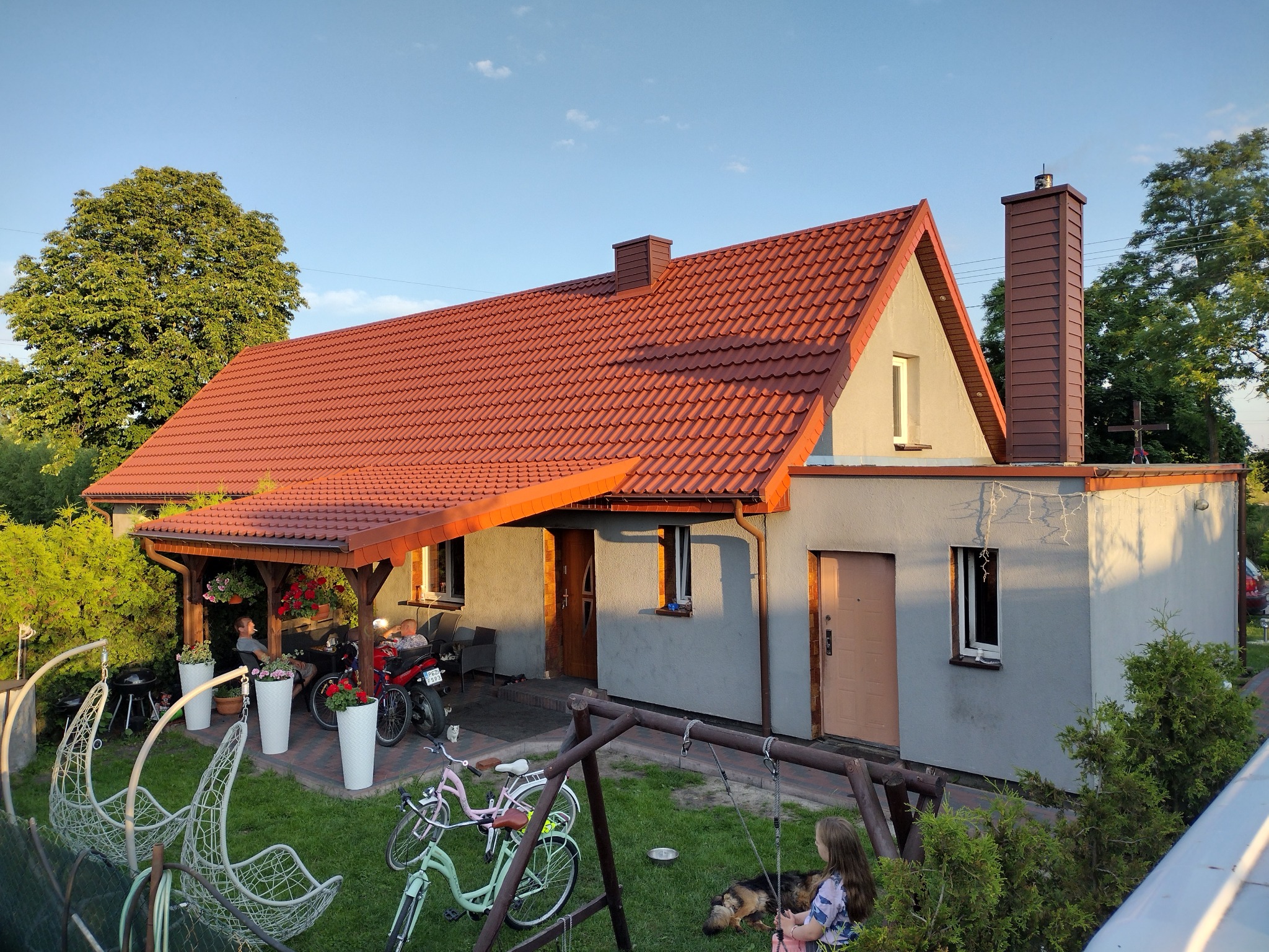 Dom jednorodzinny z czerwoną dachówką, widoczne ocieplenie poddasza od zewnątrz, z przylegającą altaną, huśtawką i rowerami na trawniku.