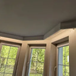 Wnętrze z trzema oknami z widokiem na zieleń, dekoracyjny sufit z ukrytym oświetleniem LED i listwami przysufitowymi.