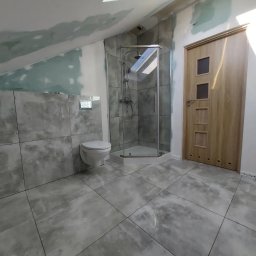 Remonty z klasą Grzegorz Dworecki - Szara łazienka na poddaszu ze szklaną kabiną prysznicową, podwieszaną toaletą i drewnianymi drzwiami z trzema kwadratowymi przeszkleniami, częściowo wykończona, z widocznymi śladami gładzi...