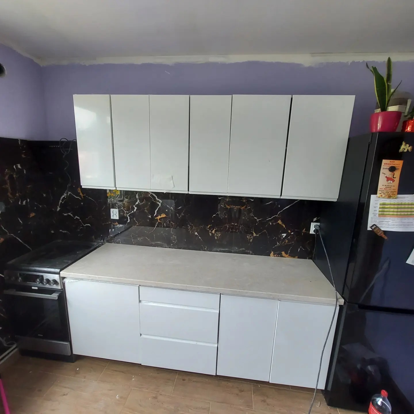 Wykończona kuchnia z białymi szafkami górnymi i dolnymi, blatem w kolorze beżowym, czarnym panelem ściennym imitującym marmur oraz sprzętem AGD w kolorze czarnym. Widoczny fragment fioletowej...