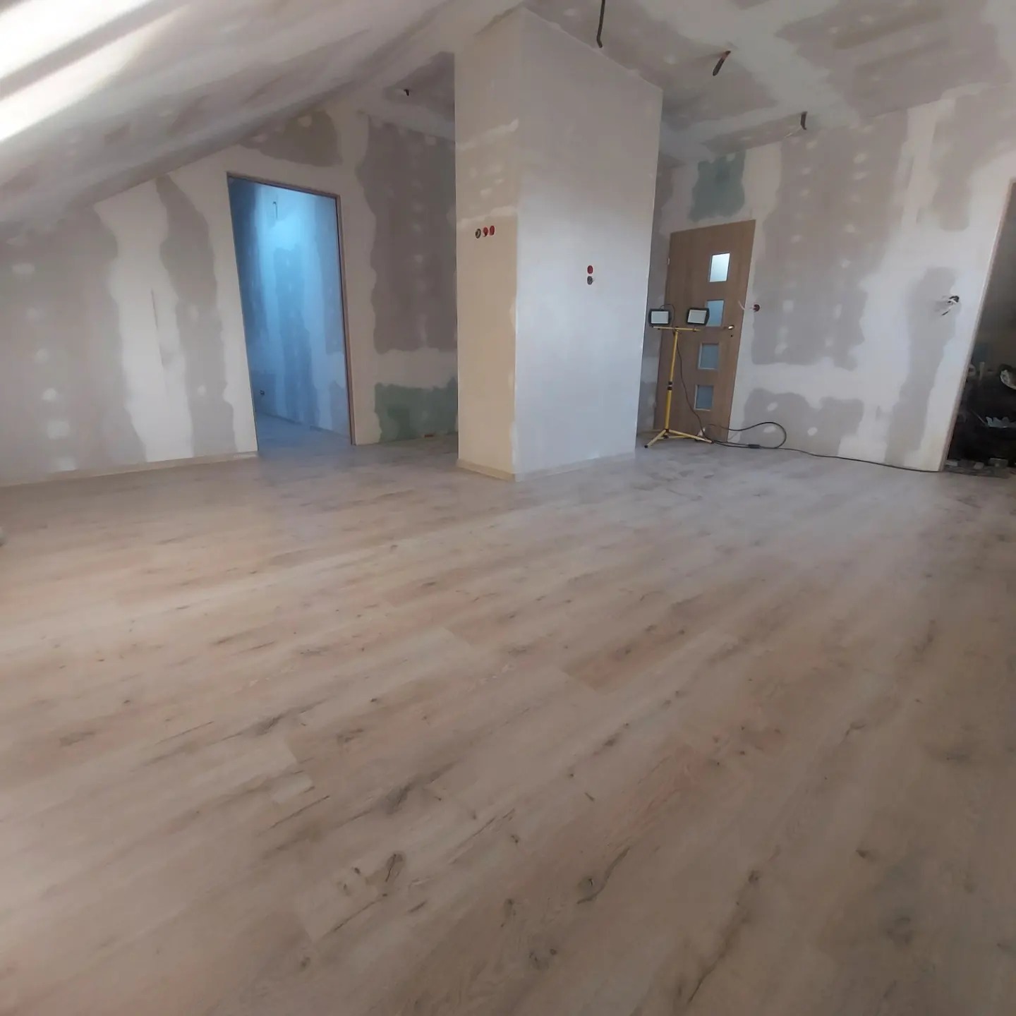 Pokój na poddaszu w trakcie remontu: położone panele podłogowe, ściany przygotowane pod malowanie, widoczne płyty karton gips i oświetlenie robocze.