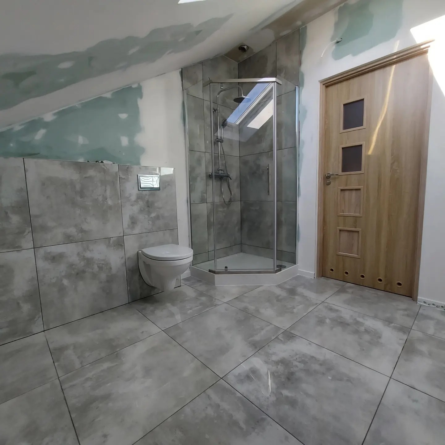 Szara łazienka na poddaszu ze szklaną kabiną prysznicową, podwieszaną toaletą i drewnianymi drzwiami z trzema kwadratowymi przeszkleniami, częściowo wykończona, z widocznymi śladami gładzi...