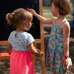 Dwie dziewczynki rysują kredą na tablicy podczas zabawy na świeżym powietrzu, widoczne białe balony w tle.