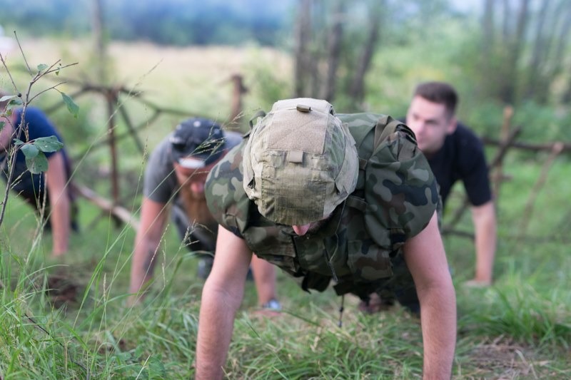 Grupa osób w strojach wojskowych wykonuje pompki na trawie w plenerze, widoczne przeszkody terenowe.