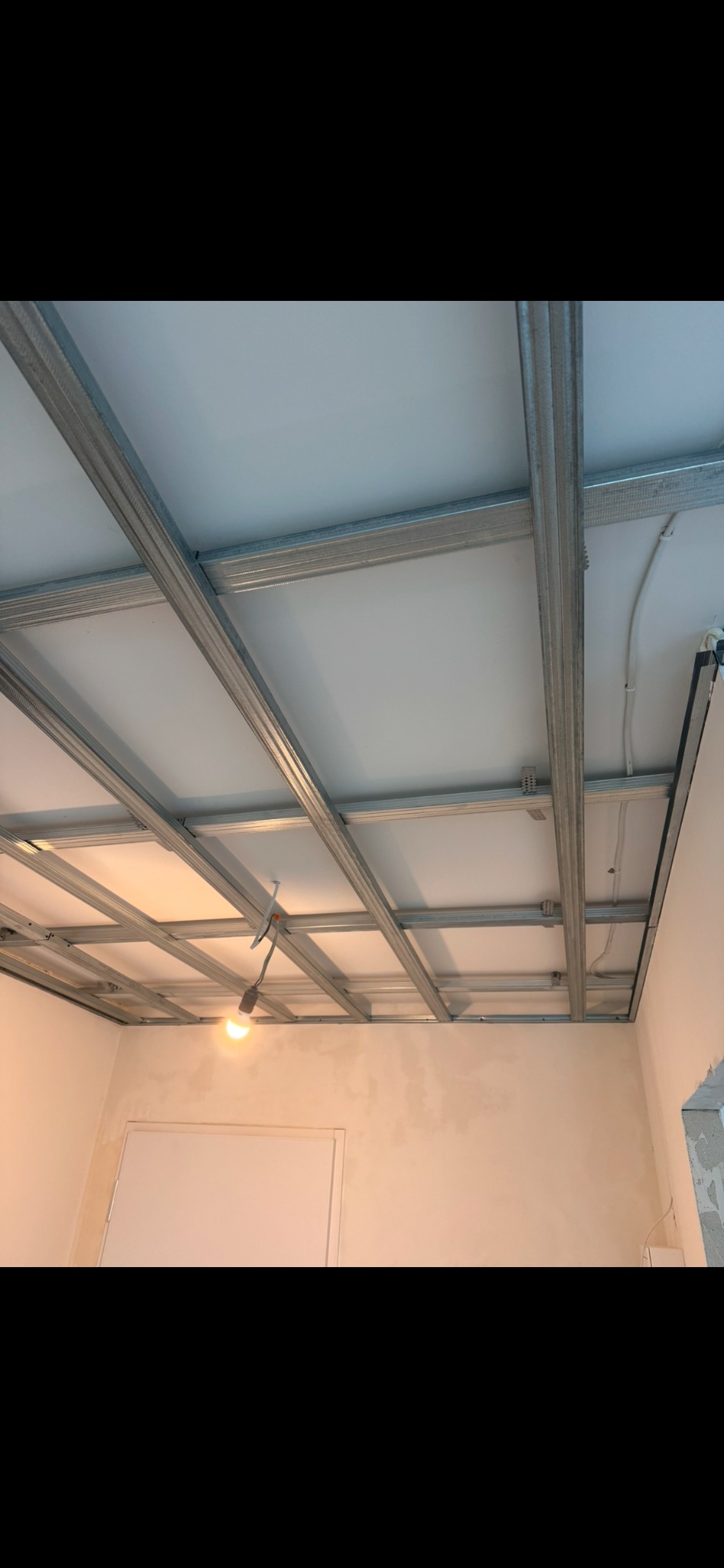 Sufit podwieszany z metalową konstrukcją i białymi panelami, widok z dołu, w tle fragment ściany i drzwi. Widoczna lampa wisząca na kablu.