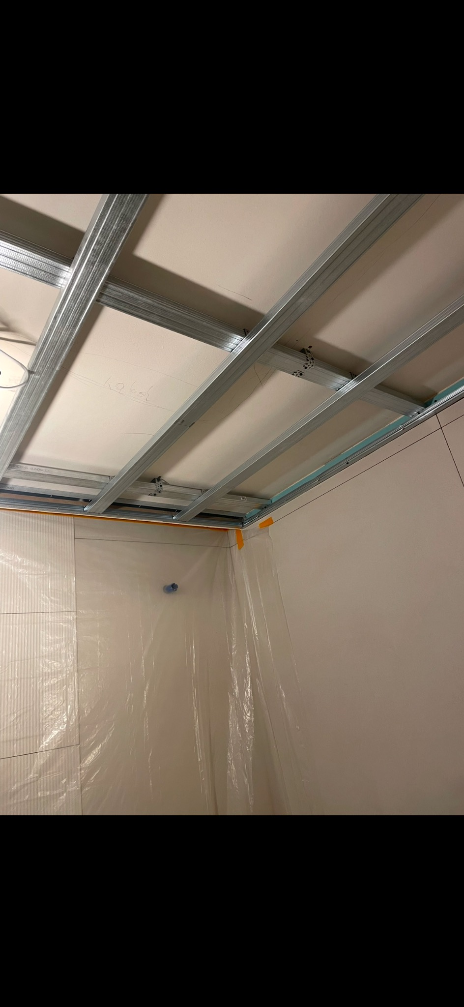 Montaż sufitu podwieszanego z metalową konstrukcją i płytami gipsowymi w pomieszczeniu zabezpieczonym folią malarską. Widoczne profile i płyty GK.