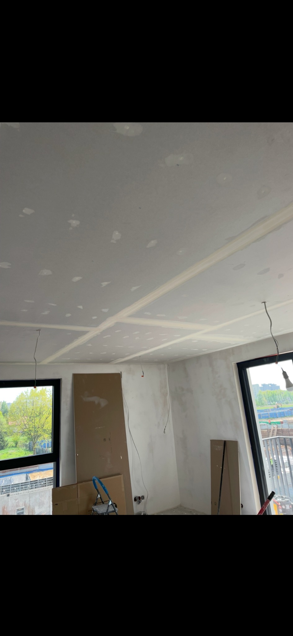 Wnętrze w trakcie prac wykończeniowych: szpachlowana sufit i ściany, widoczne okna z widokiem na zieleń, kartony i drabina. Etap przygotowania do malowania.