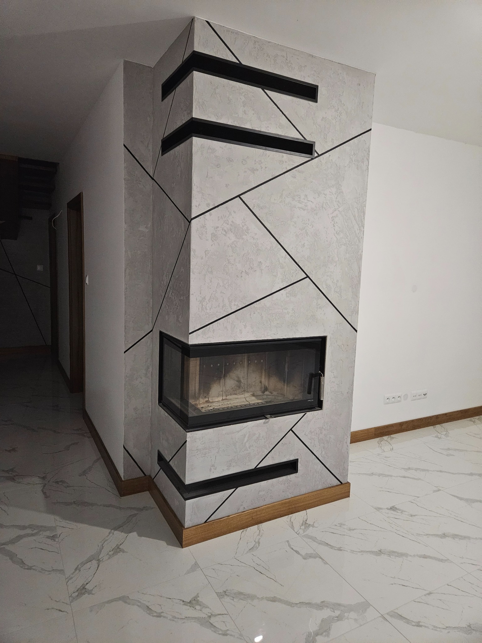 Wykończony kominek z czarnymi geometrycznymi zdobieniami na szarej ścianie, obudowany drewnianą ramą, na podłodze z białego marmuru.