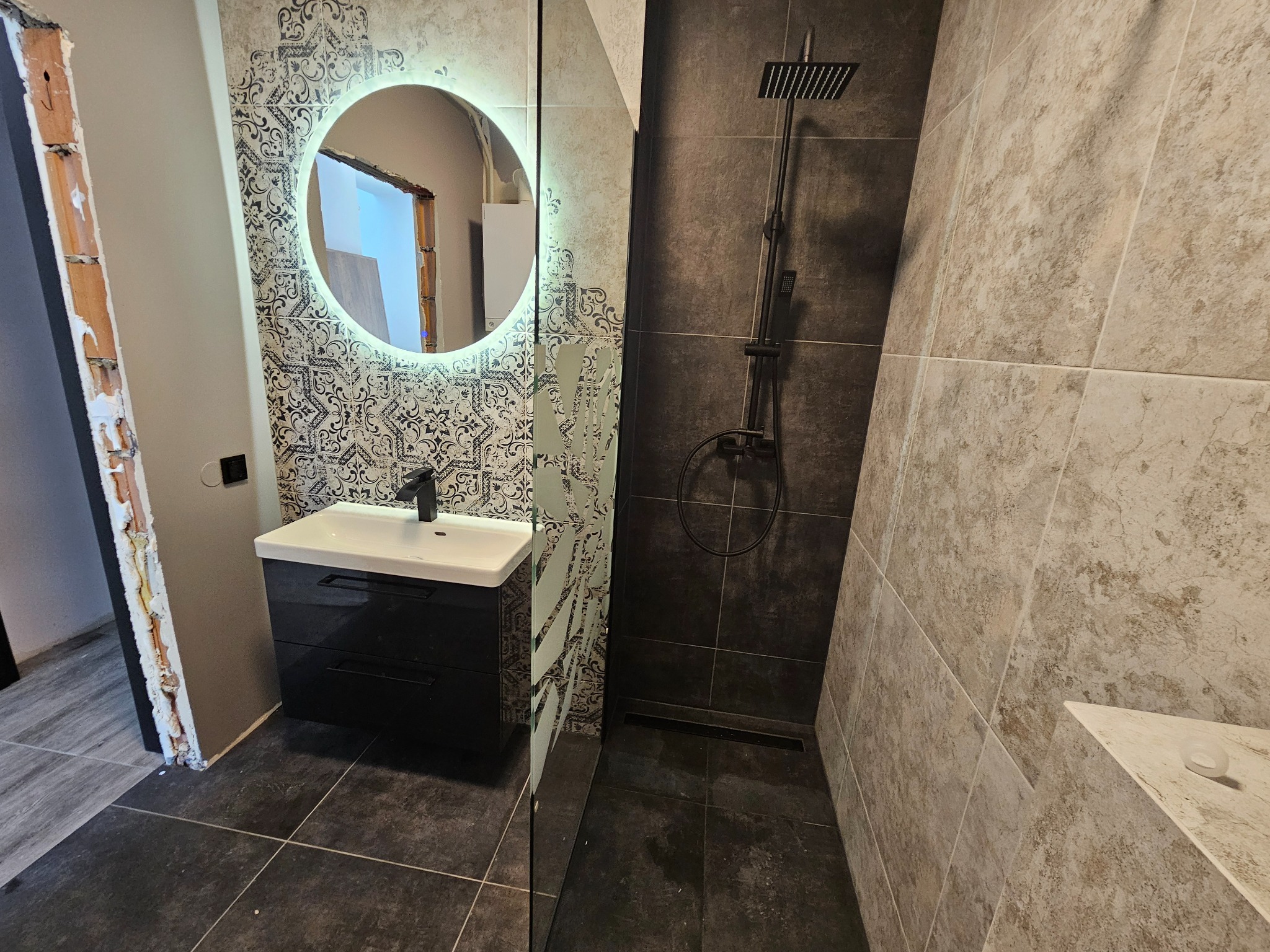 Nowoczesna łazienka z czarną armaturą, okrągłym lustrem z podświetleniem LED, czarną szafką pod umywalką i kabiną prysznicową typu walk-in, wykończona płytkami w odcieniach szarości i beżu.