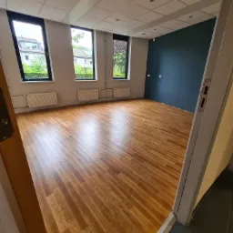 Pusta przestrzeń z ułożoną drewnianą podłogą, widok zza otwartych drzwi, trzy okna z widokiem na zieleń, jedna ściana pomalowana na ciemny niebieski kolor.