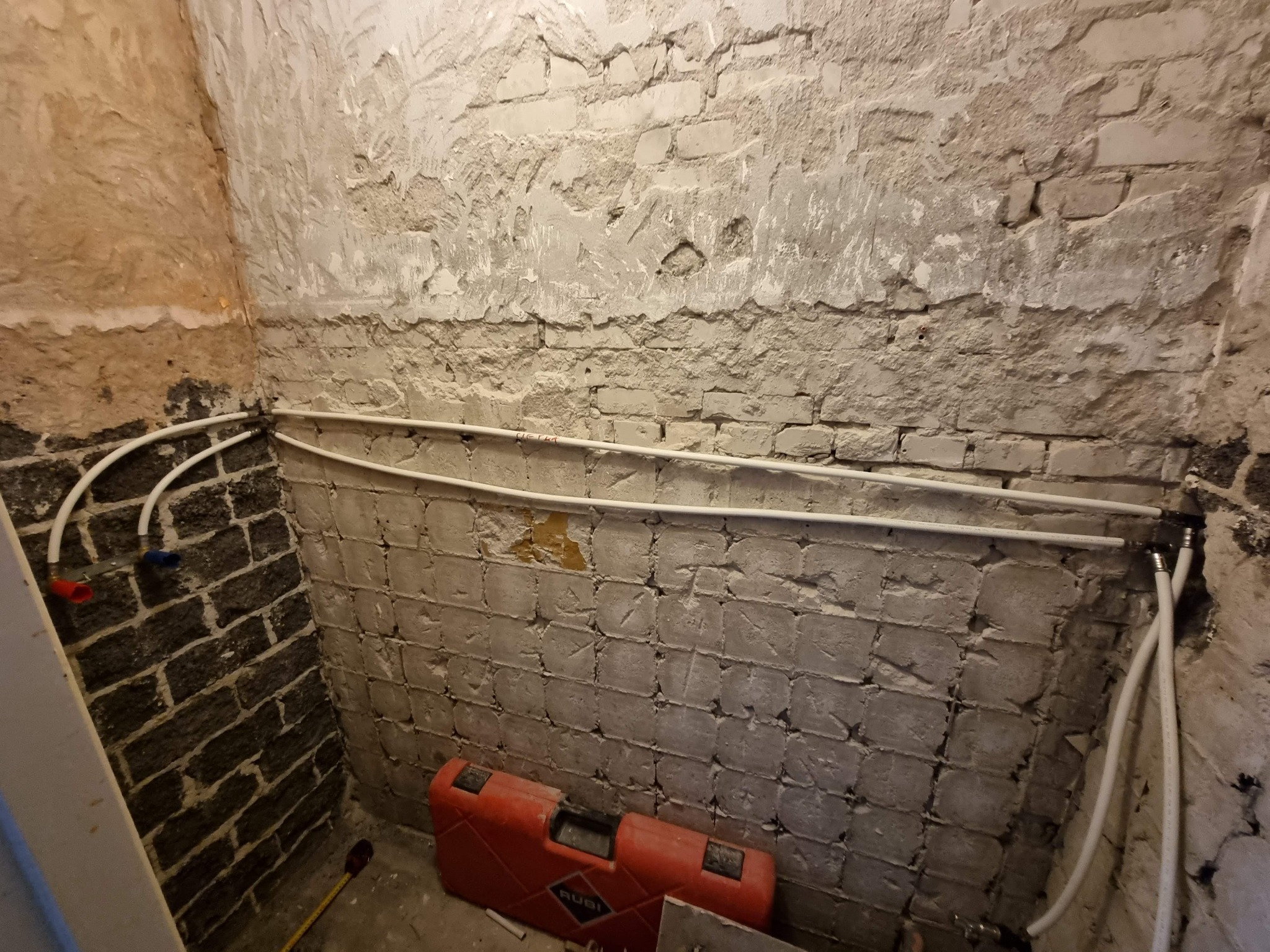 Surowa ściana w łazience z widocznymi rurami instalacji wodnej, czerwonym i niebieskim zaworem oraz narzędziem glazurniczym marki Rubi.