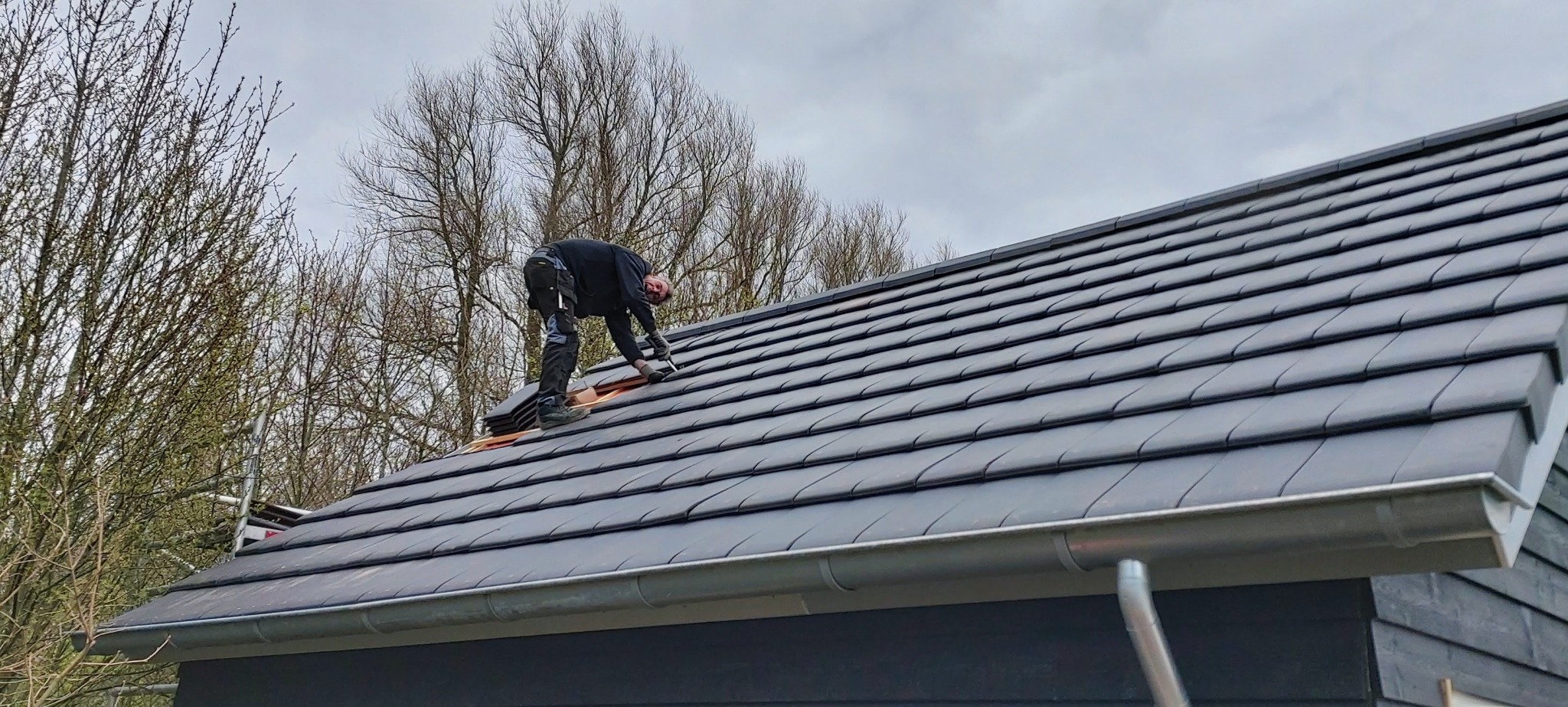 Pracownik w czarnym ubraniu układa grafitową dachówkę na dachu budynku, na tle drzew bez liści i pochmurnego nieba.