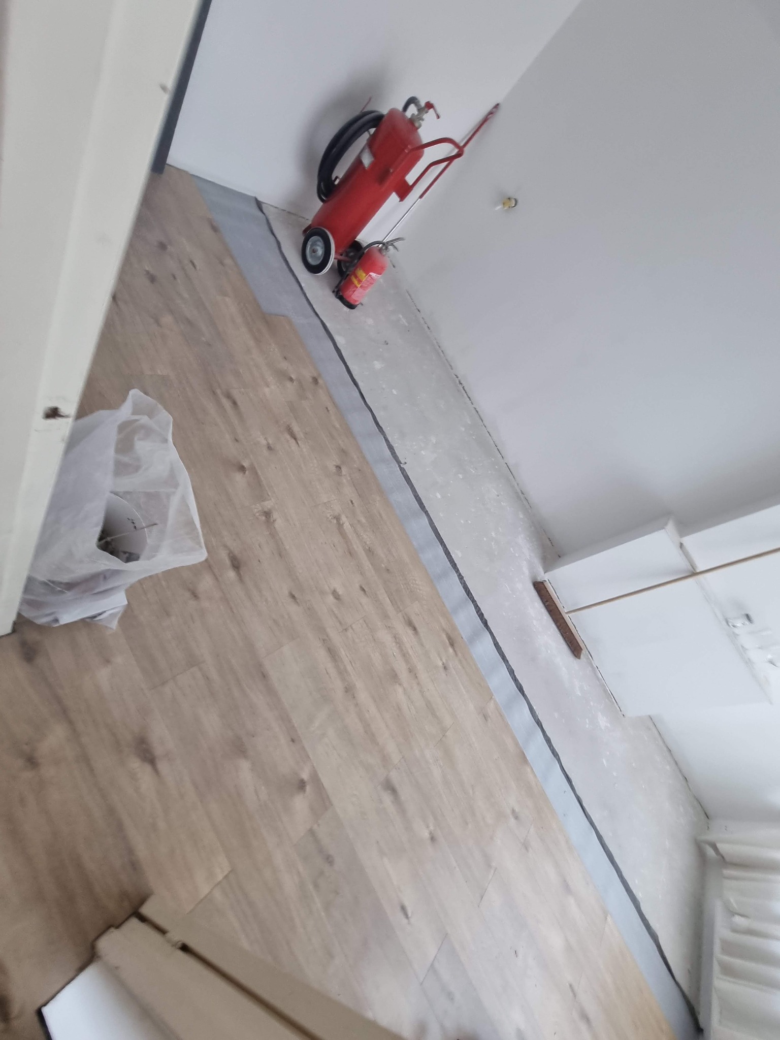 Fragment pokoju w trakcie układania paneli podłogowych, widoczny czerwony wózek z gaśnicami, podkład pod panele i częściowo ułożone panele z jasnego drewna.
