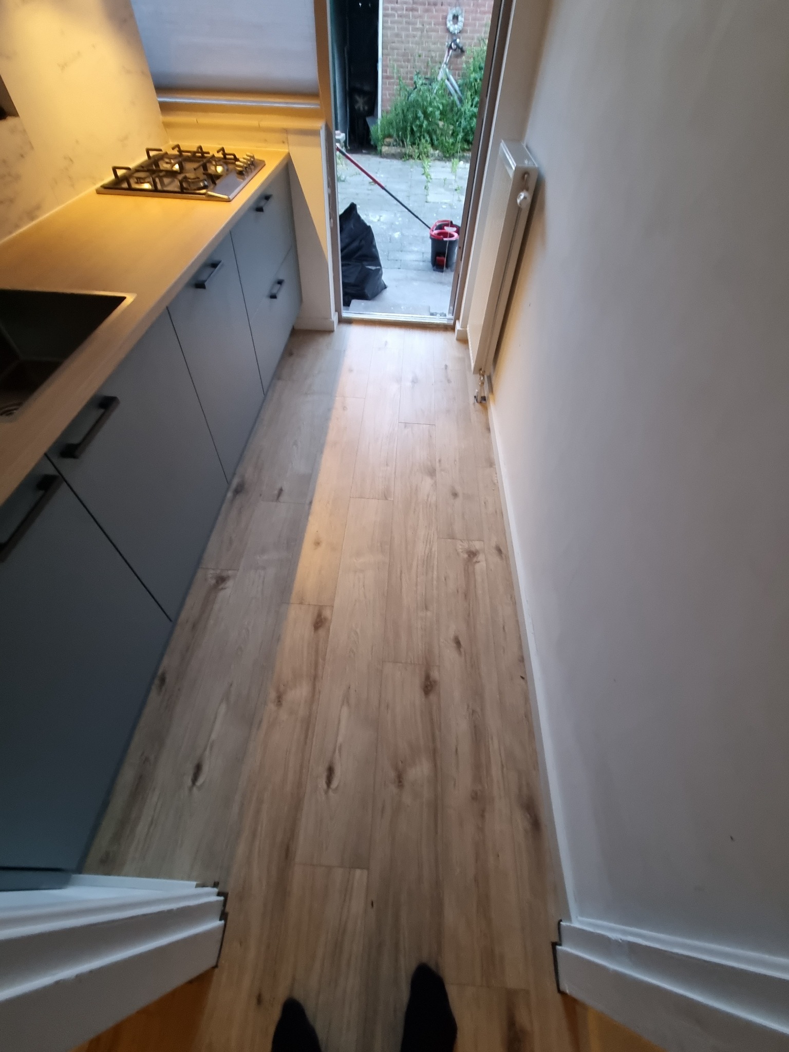 Nowoczesna kuchnia z szarymi szafkami i blatem w odcieniach beżu, z widoczną płytą gazową, jasnymi panelami podłogowymi i fragmentem grzejnika przy białej ścianie. Widok w kierunku otwartych drzwi...