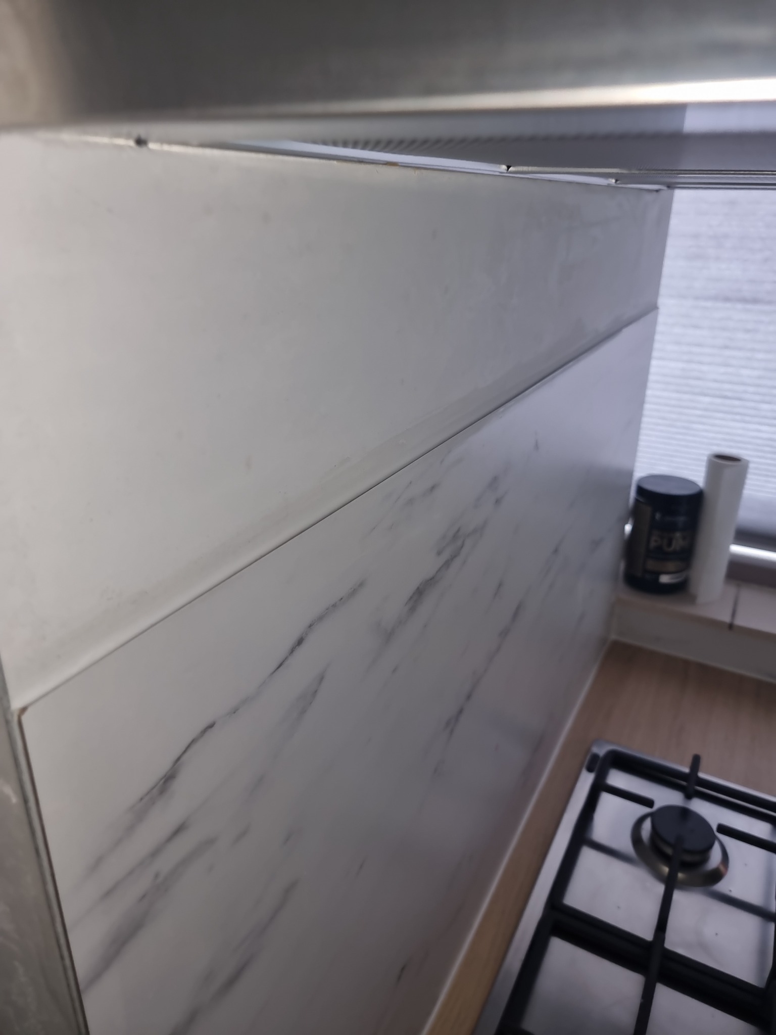 Fragment ściany nad kuchenką gazową, wykończonej białymi panelami imitującymi marmur, z widocznymi elementami okapu i blatem. Na parapecie okiennym stoi czarny pojemnik i rolka papieru.