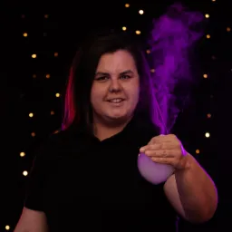 Kobieta uśmiecha się, trzymając w dłoni przezroczystą kulę z fioletową parą na tle ciemnej ściany z lampkami.